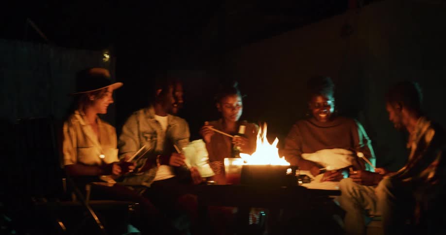 A group of men and women sit around a fire roasting marshmallows and laughing