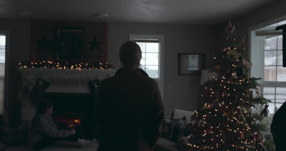 Older woman walks into room past christmas tree toward teenage girl sitting on floor near fireplace