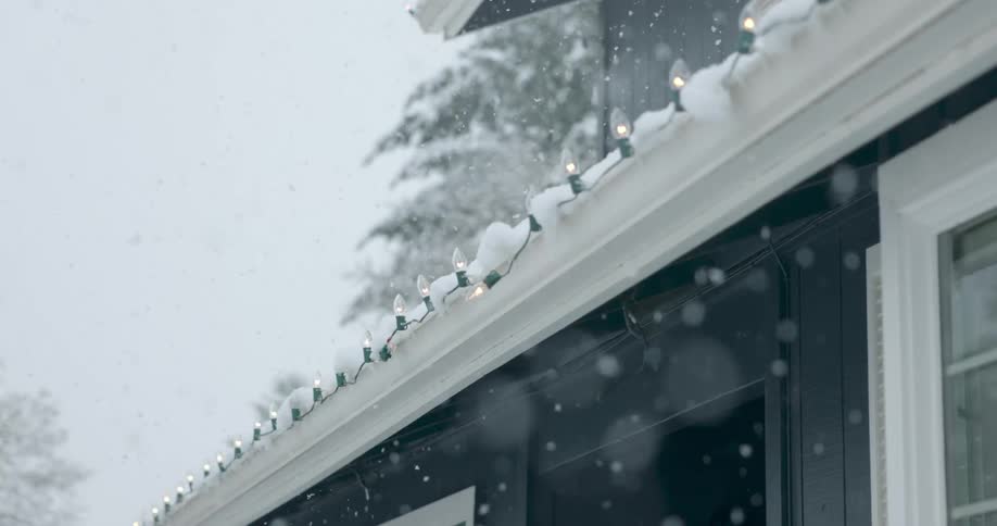 Christmas lights on a house shine as snow gently falls from the winter sky