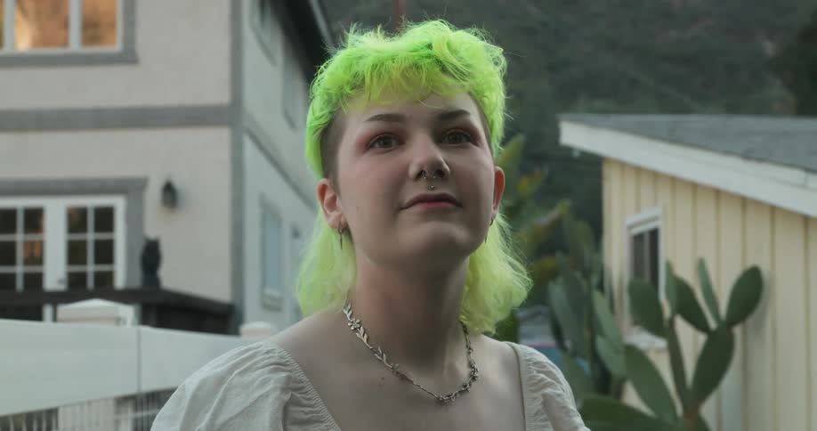 A teenage girl with bright green hair is standing outside in a neighborhood looking around