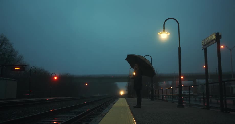 Woman with black umbrella coat and pants stands in train station as it rains