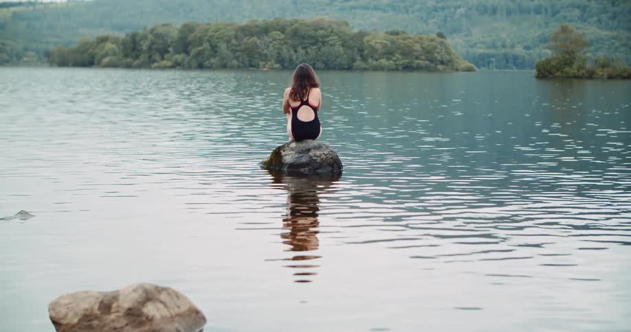 Woman with wavy hair and black swimsuit sits on rock watching lake
