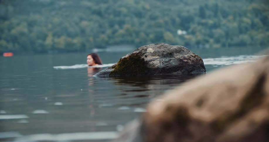 Woman with red hair swims in lake next to mountain with forest