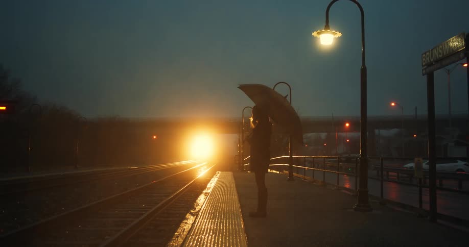 Person with umbrella waits near tracks silhouetted by light of approaching train at night