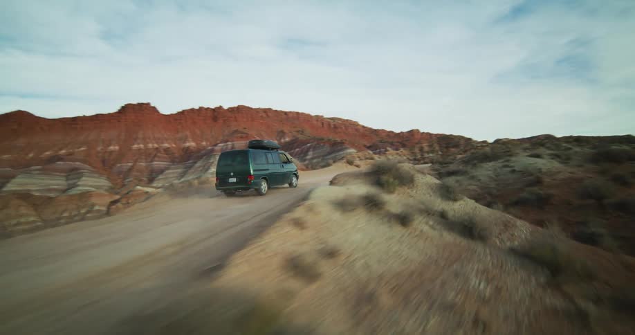 Vehicle drives along dirt road through badlands under a cloudy sky
