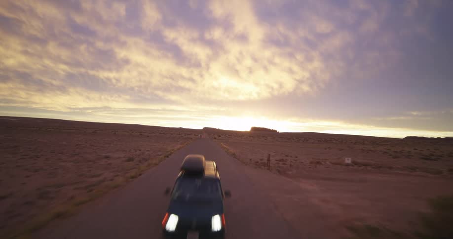The headlights on a van shine as it travels on a desert road with bright clouds in the sky
