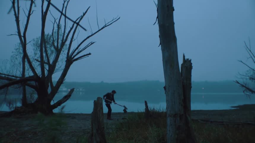 A man digs a hole in a wooded area by a body of water