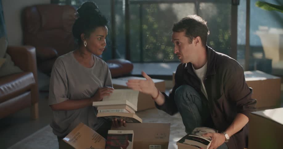 Man and woman go through books in one of several cardboard boxes sitting on living room floor
