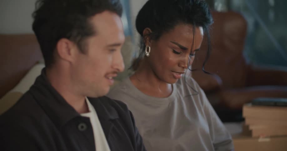 Woman with gray t shirt green pants white shoes sits on sofa with man wearing shirt and jeans using laptop in living room at home