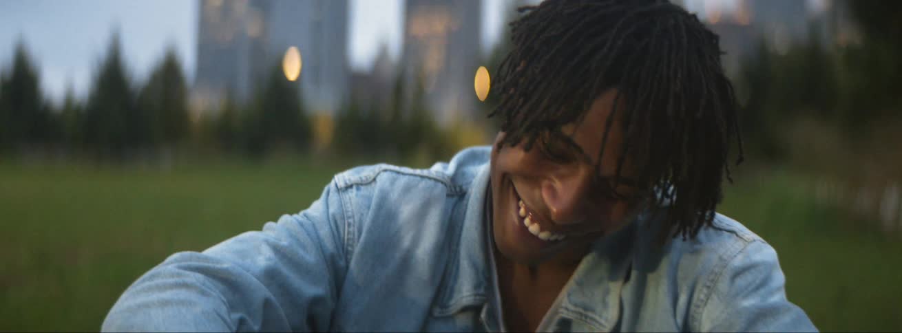 Man sits in field laughing with someone else with city skyline in the background