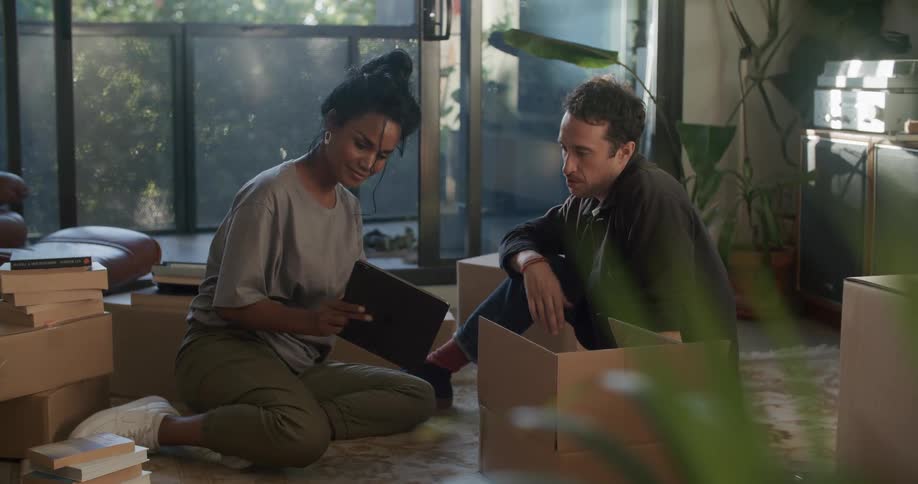Woman with gray t shirt green pants white shoes sits on rug with man wearing shirt and jeans looking at tablet in living room at home