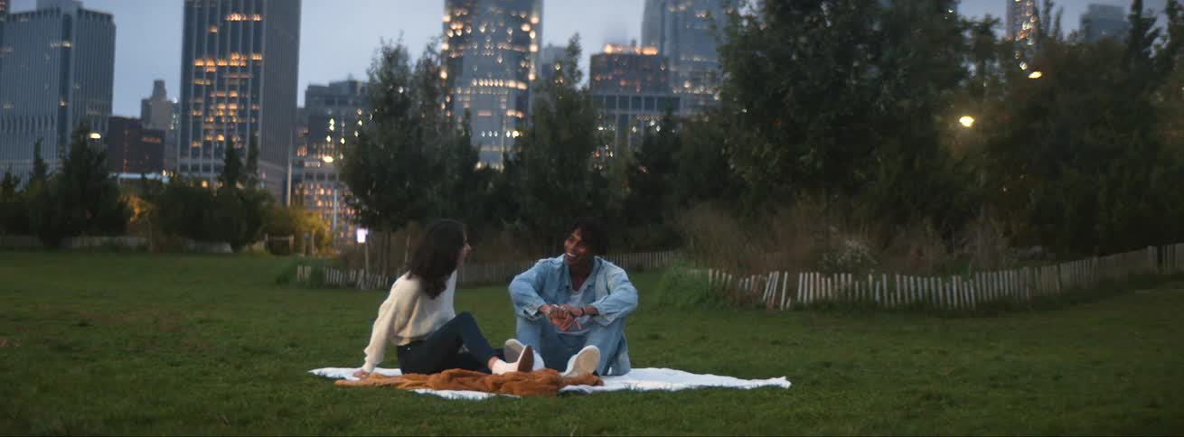 Young couple sitting over a blanket in a park laughing and smiling in front of the city skyline