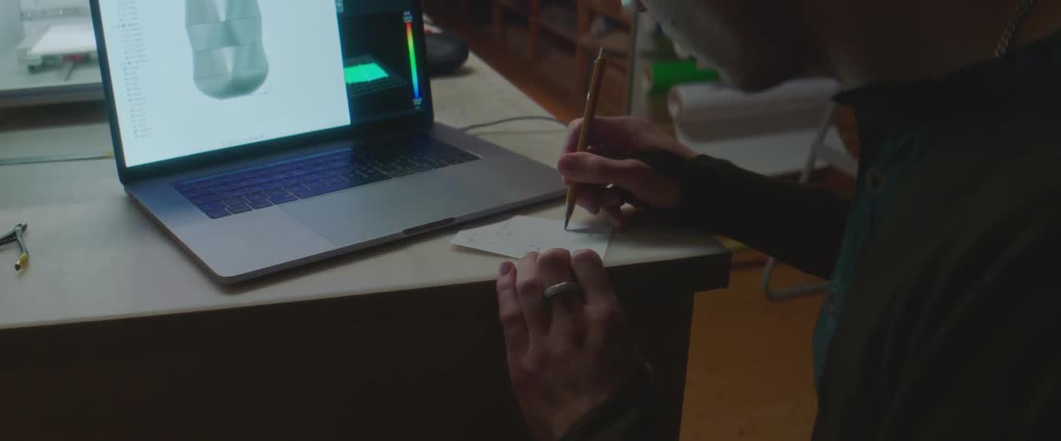 Young man writing with a pencil on a small piece of paper over a desk in front of a laptop