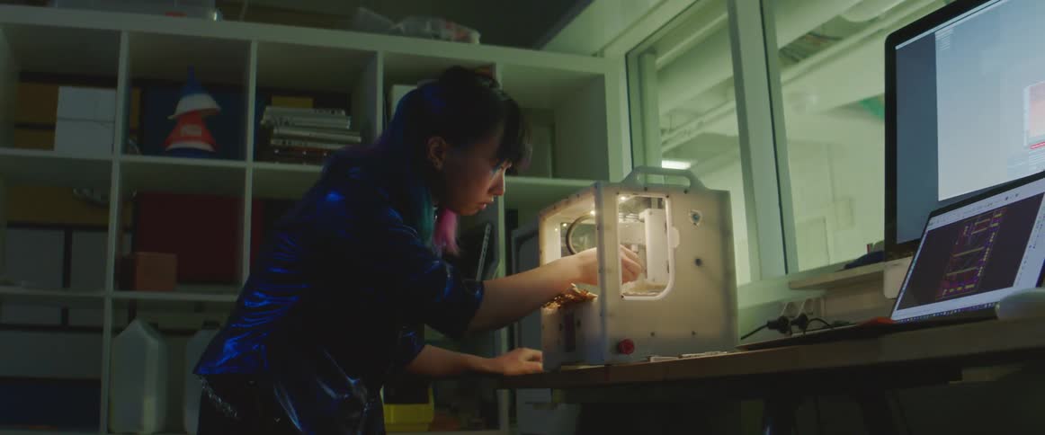 An asian woman is working with a 3d printer and a laptop computer is on the desk