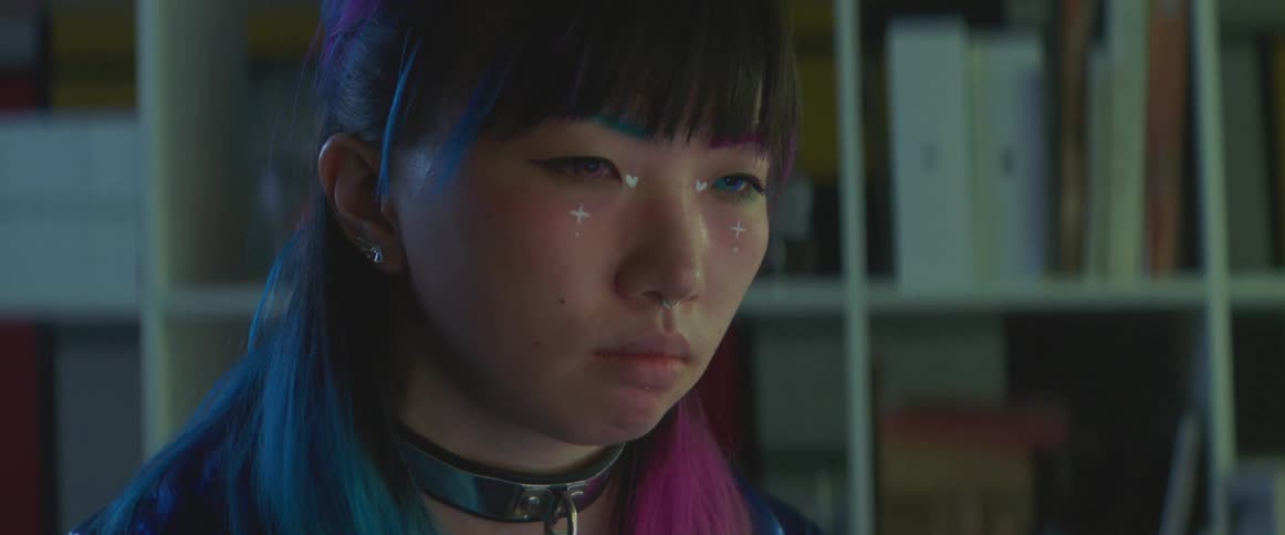 Teenage girl with colorfully dyed hair face paint and colored contacts sits near shelves