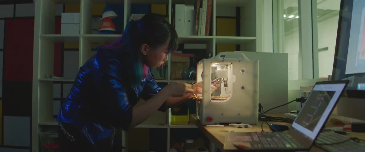 A woman is leaning over to fix a 3d printer in a room full of electronics