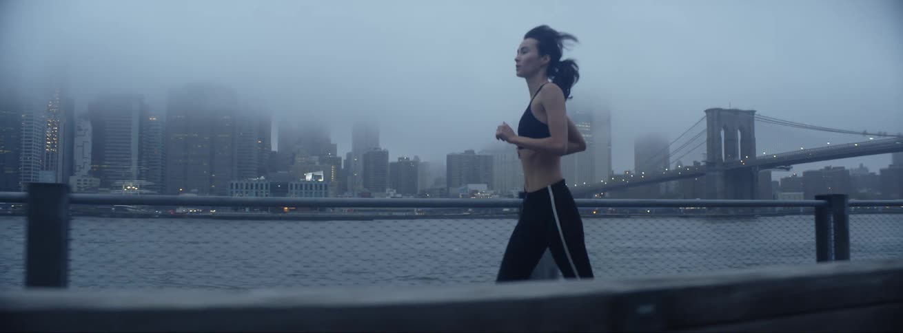 Woman with sports bra and leggings runs along promenade with high rise buildings and brooklyn bridge on cloudy day