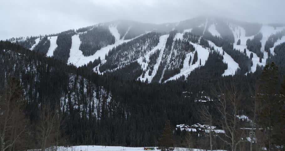 A white truck is traveling on a snow covered mountain forest road with a snow covered mountain range in the distance under a cloudy sky
