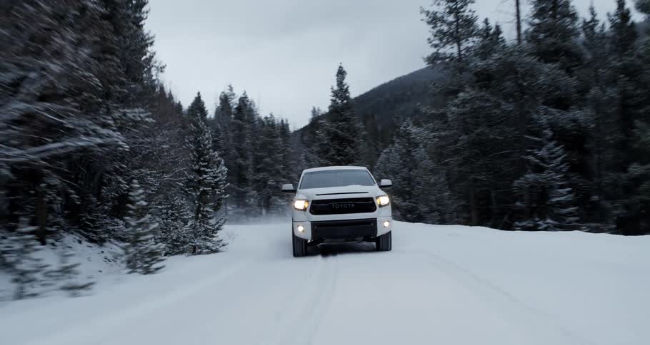 A white truck is traveling on a snow covered forest mountain road with a mountain range in the distance