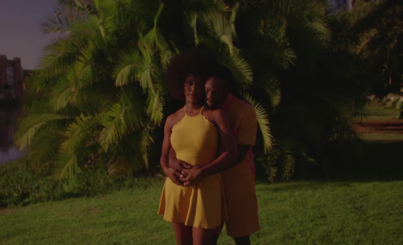 A man hugs his girlfriend from behind as they pose in a tropical field