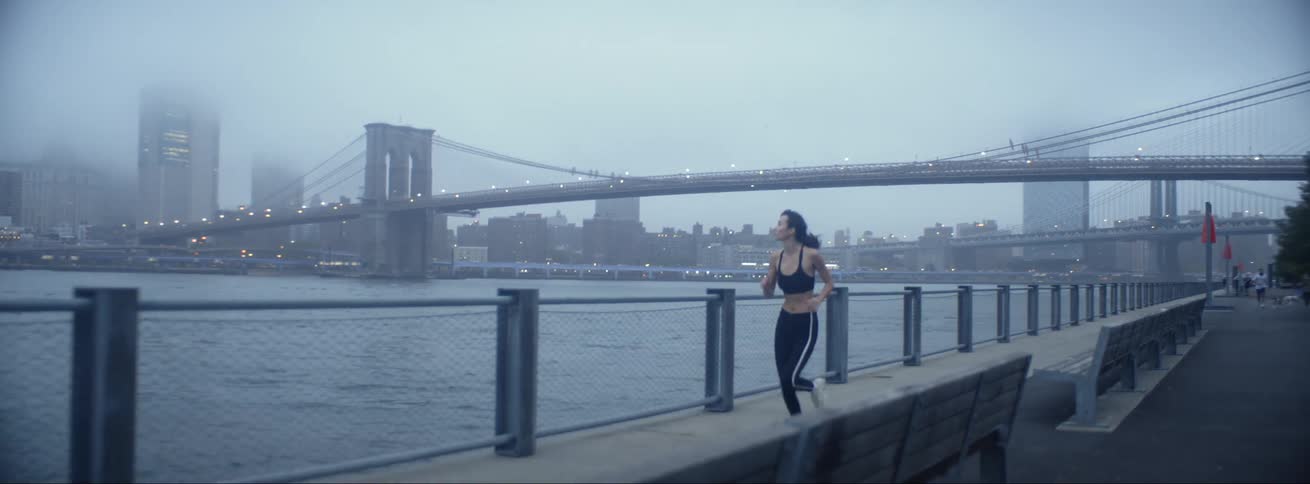 Woman with sports bra and leggings runs along promenade with high rise buildings and brooklyn bridge on cloudy day