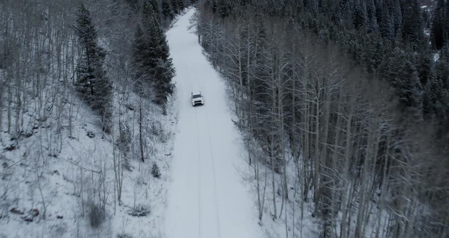Aerial view of a white truck with the headlights on traveling on a snow covered forest mountain road