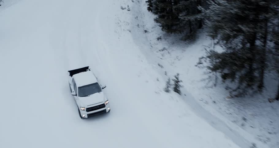 Aerial view of a white truck traveling on a snow covered forest road lined with snow covered trees