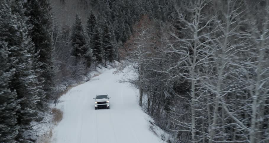 Aerial view of a white truck with the headlights on traveling on a snow covered forest mountain road