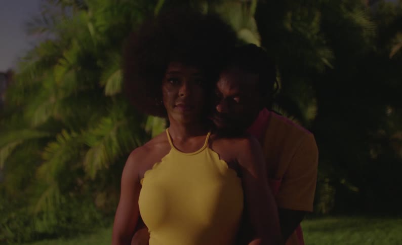 Female dancer with curly hair and yellow dress and male dancer with braids and pink yellow shirt standing in front of trees look at camera smiling in public park