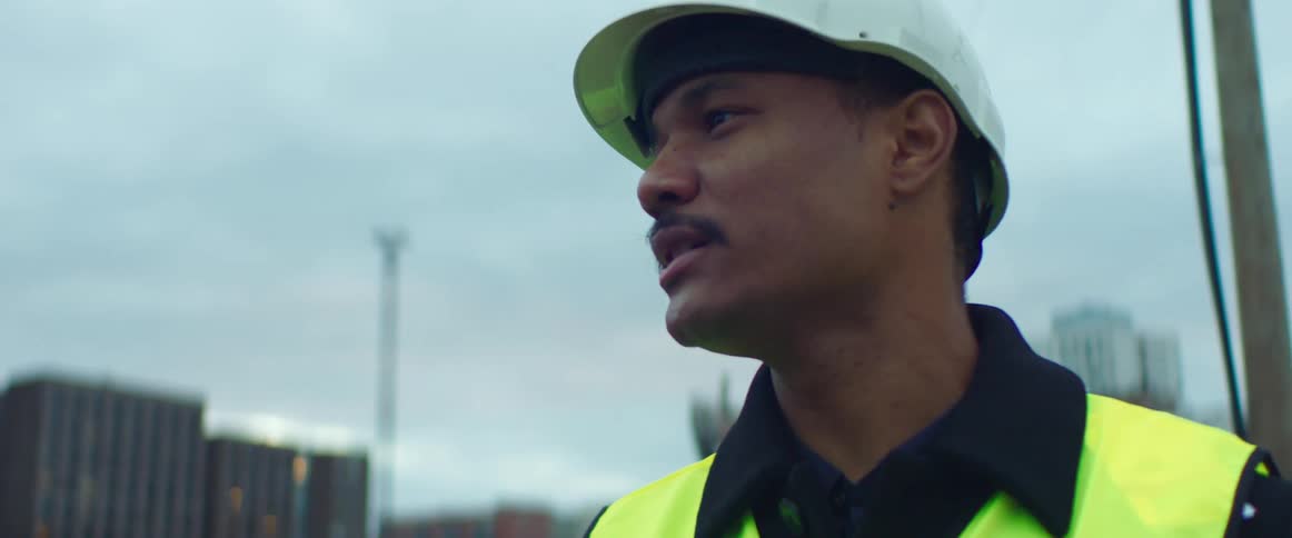 A man wearing a hardhat and safety vest talks with city buildings and equipment behind him
