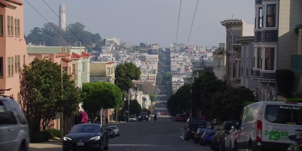 Cars travel on a hilly neighborhood street with a tower on the skyline