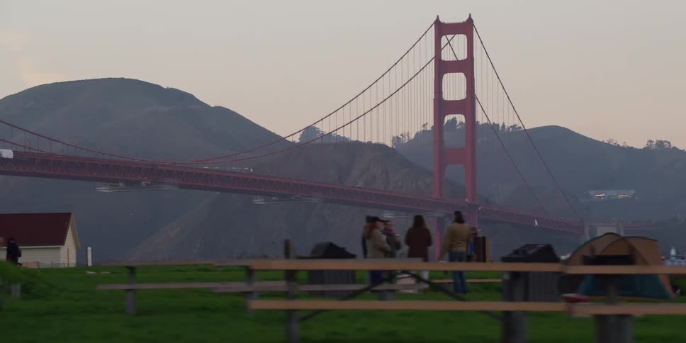 Distant view of suspension bridge with suspension cables on vertical suspenders and public park with people at dusk