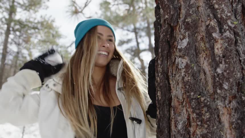 A laughing woman throwing a snowball before ducking behind a tree