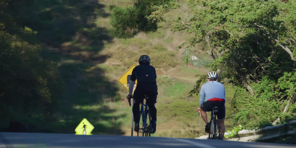 Two male cyclists with helmets and jerseys cycle downhill on highway