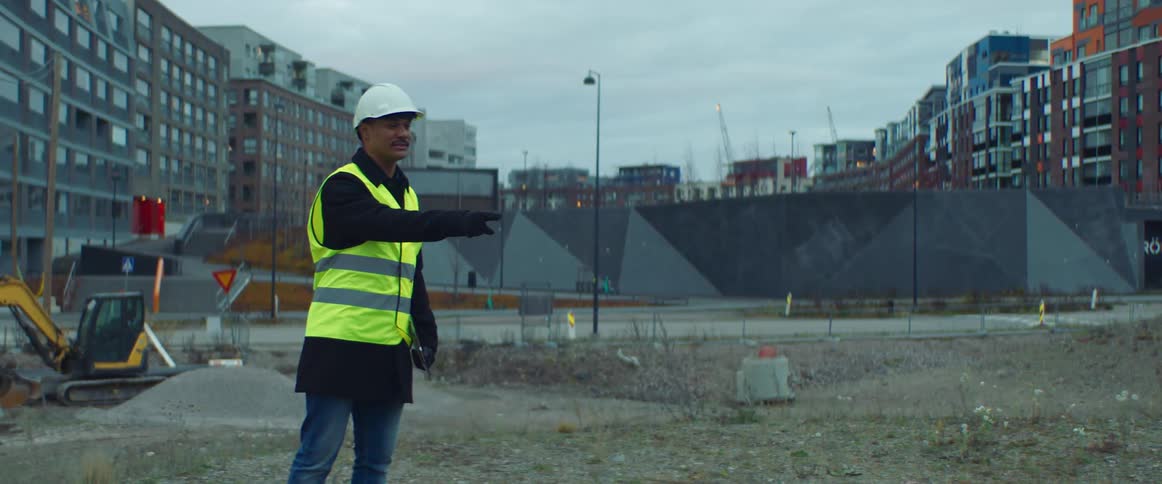 A construction worker stands in a construction site in front of a bulldozer and puts his arm out as he tells someone to stop