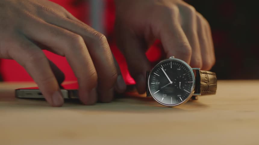 Mans hand picking up his smartphone and wristwatch from a wooden table
