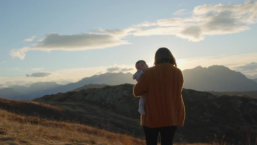 Mother holding a baby in her arms standing on a hill near the mountains at sunset