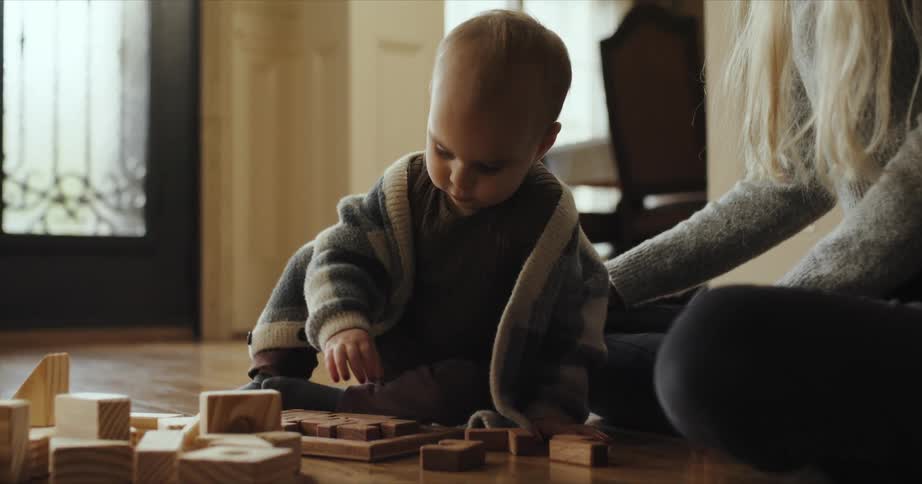 A woman watches as her baby plays with wooden blocks on the floor