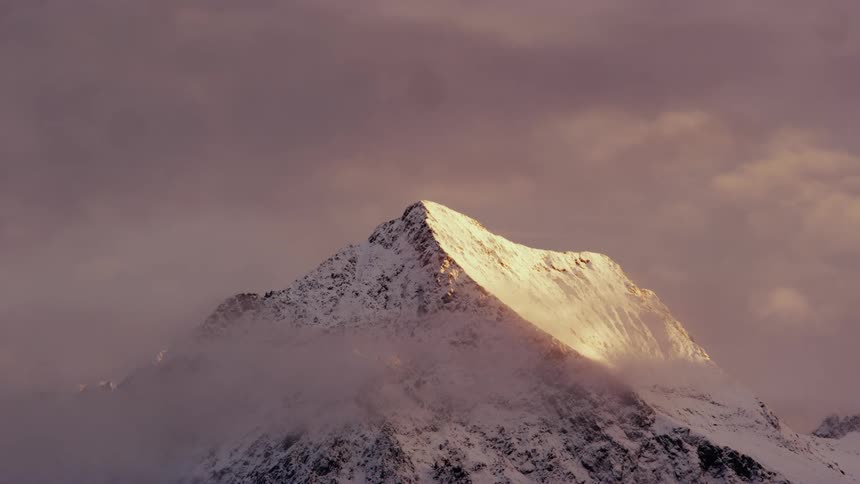 Sunlight shines on a snow covered mountain peak high in a pink cloudy sky