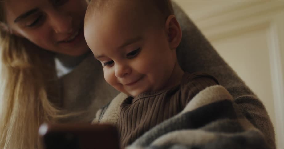 Blonde mother with gray sweater holds toddler boy using smartphone