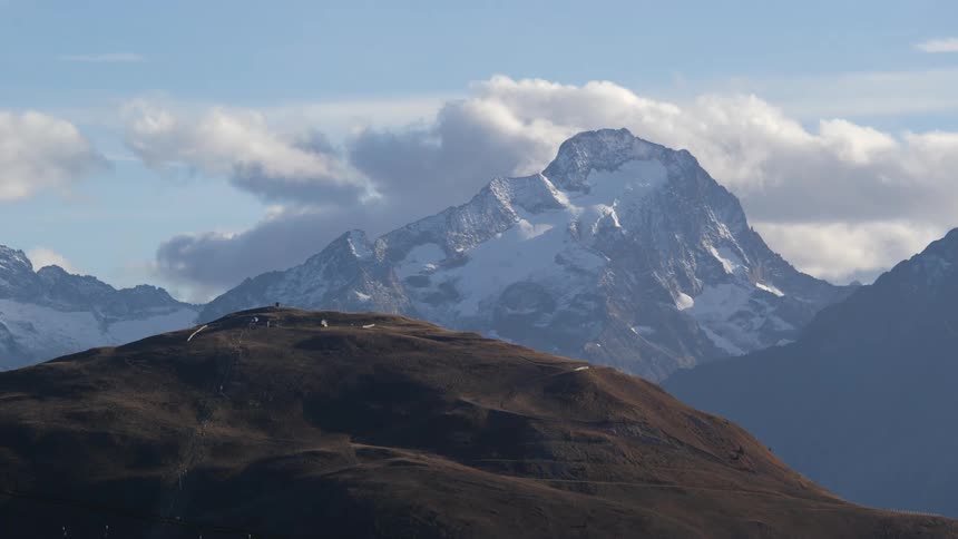 A dirt covered mountain top with a snow covered mountain ridge in the distance among the clouds