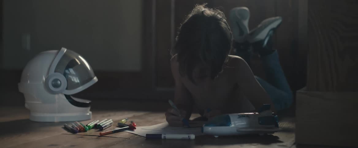 A boy lying at the floor and drawing on a paper next to his spaceship and space helmet toys