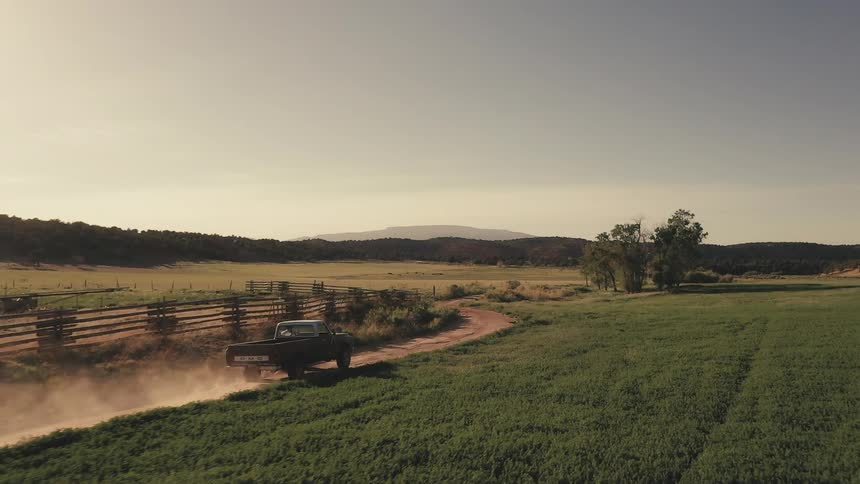 A pickup truck travels on a rural dirt road with mountains on the horizon