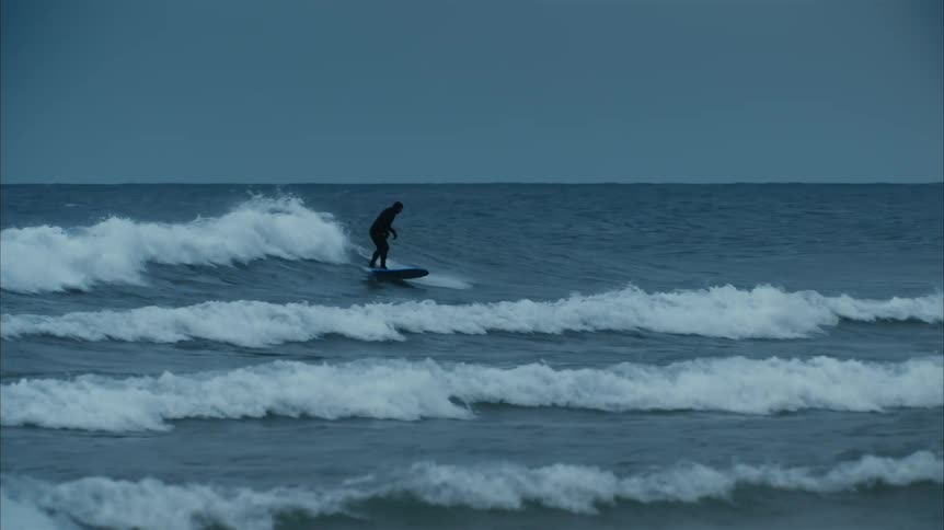 Surfer wearing a full body black wet suit rides a blue surfboard on the ocean waves close to the coast