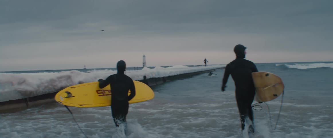 Wet suit wearing surfers carry surfboards into ocean near snow covered pier
