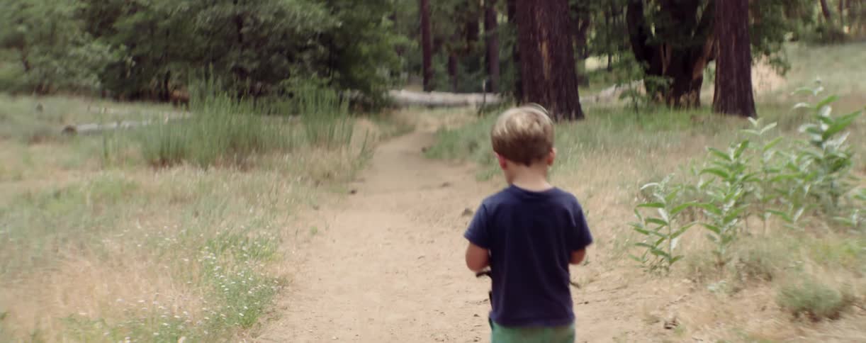 A toddler boy is walking alone on a dirt path in a wooded area