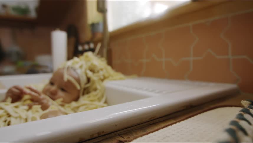 Baby reclines in kitchen sink covered in noodles eating as person piles noodles onto their head