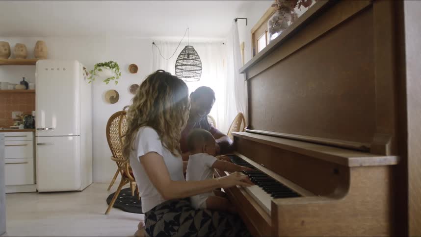 Mother with brown wavy hair and white shirt holds baby son and playing piano next to father with long hair and red shirt at home