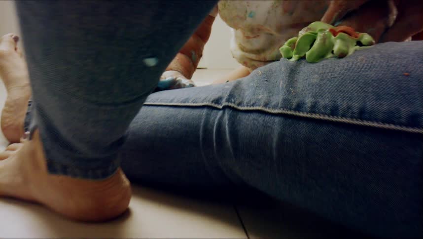 A woman is sitting on the kitchen floor while her gloved toddler is putting paint on her jeans