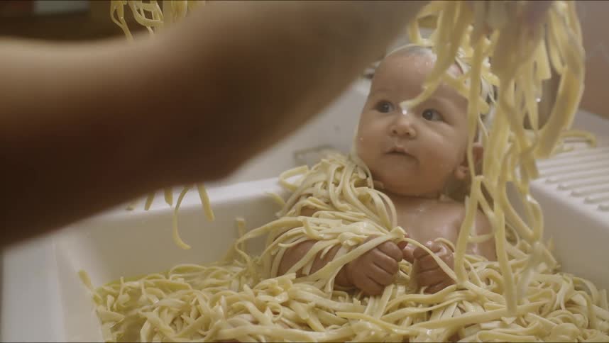 A baby is sitting in a bathtub with raw noodles while a woman plays with her
